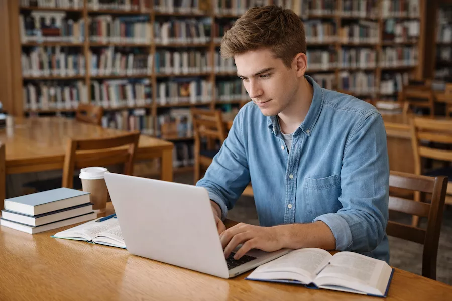 Student som pluggar i bibliotek med böcker och laptop – symboliserar studiero, fokus och effektivt lärande i akademisk miljö.